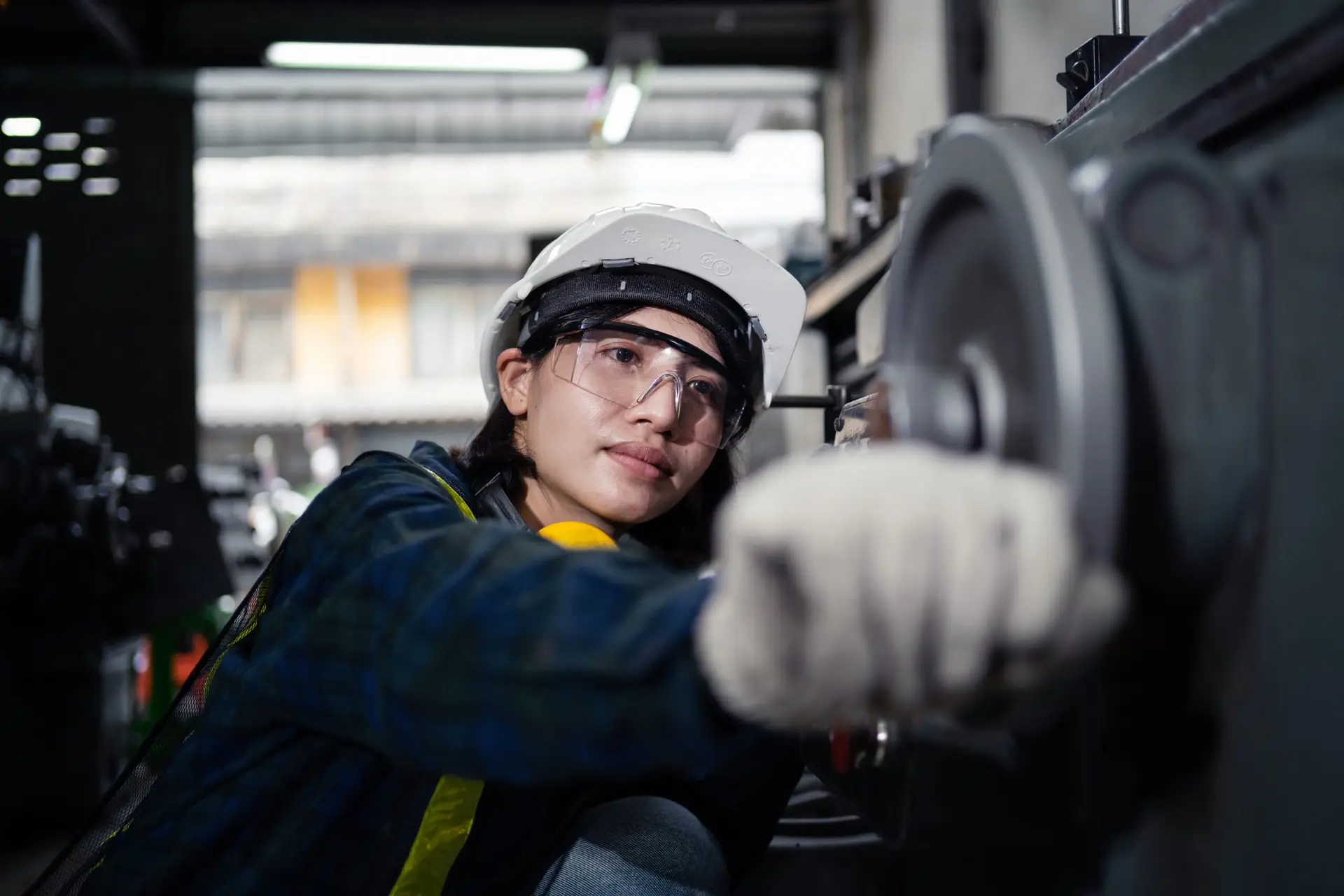 A woman wearing a yellow helmet and safety gear is pointing at a piece of machinery. She is likely a worker in a factory or industrial setting